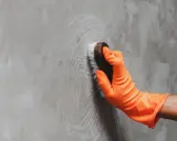Mano con guante naranja limpiando pared gris con cepillo, aprendiendo como limpiar las paredes de color sin dañar la pintura.