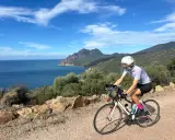 Ciclista pedala lungo la costa rocciosa della Corsica, con il mare blu e montagne sullo sfondo. Un'avventura in bici indimenticabile.