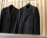 Dwa eleganckie garnitury wiszą na wieszakach. Jeden w prążki, drugi z subtelnym wzorem. Prasowanie garnituru to klucz do nienagannego wyglądu.