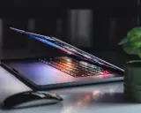 Otwarty notebook z podświetlaną klawiaturą i ekranem, obok myszka i roślina. To jest właśnie notebook, narzędzie do pracy i rozrywki.