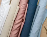 Rullar av tyg i olika färger, från vitt till ljusblått, med en rosa och beige nyans emellan. Bomullsegenskaper gör dessa tyger mångsidiga för sömnad.