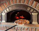Une pizza garnie est sur le point d'entrer dans un four à pizza extérieur en briques rouges, avec des flammes visibles à l'intérieur.