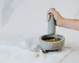Dłoń uciera w moździerzu aromatyczne pesto z bazylii. Obok leżą ząbki czosnku.