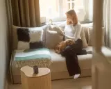 En kvinna läser en bok i en liten bäddfåtölj med sin hund i knät. En grön och vit pläd ligger över fåtöljen.