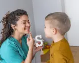 Logopeda pomaga dziecku z dyslalią ćwiczyć wymowę litery "S", używając białej literki.