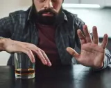 Mężczyzna z zarostem odpycha szklankę z alkoholem, walcząc z chorobą alkoholową. Gest dłoni symbolizuje odmowę i chęć zmiany.