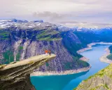 Två personer står på Trolltunga, en av Norges mest ikoniska och fina platser. En dramatisk klippavsats med en turkos sjö nedanför och snöklädda berg i bakgrunden.