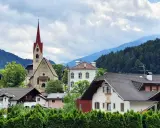 Un borgo pittoresco in Val d'Isarco, con una chiesa dal campanile rosso svettante tra case tradizionali e montagne boscose.