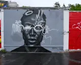 En imponerande **graffiti** av ett ansikte med en krona och solstrålar runt ögonen. Konstverket är målat i gråskala med vita detaljer.