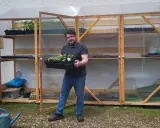 Un homme souriant tient un plateau de jeunes plants devant sa serre pour semis fait maison, remplie d'étagères de semis.