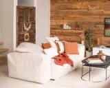Un salon chaleureux et accueillant avec un grand canapé blanc, des coussins orange et un mur en bois rustique. Une porte ancienne décore le mur.