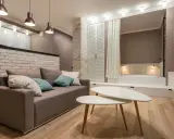 Nowoczesny salon z sofą i stolikami kawowymi. W tle widoczna ściana ze starej cegły, łóżko i szafa.
