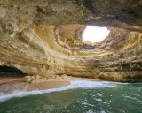 Die Benagil-Höhle mit ihrem kreisförmigen Loch im Felsdach, das Sonnenlicht hereinlässt. Wellen brechen sanft am Sandstrand im Inneren.