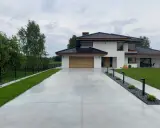 Nowoczesny dom z drewnianą bramą garażową, otoczony zielenią. Szeroki podjazd betonowy prowadzi do wejścia, a wzdłuż niego biegną lampy i rabaty z kamieniami.