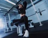 Donna in tenuta sportiva nera, con scarpe bianche, seduta su panca in palestra. Ispirazione su come vestirsi in palestra.
