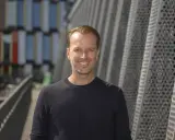 Daniel Larsson, skådespelare, ler mot kameran. Bakgrunden är en modern byggnad med geometriska mönster.