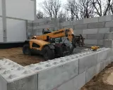 Żółty ładowacz teleskopowy Liebherr pracuje przy budowie muru oporowego z betonowych bloków.