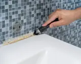 Mano usando una espátula para quitar silicona vieja y sucia de la bañera.