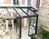 Une véranda bioclimatique moderne, avec une structure en verre et métal noir, s'intègre à une maison en briques et pierre. Avis positif sur son esthétique.