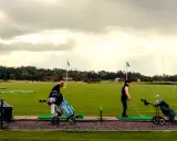 Kvinnor tränar golf på en driving range i Sverige. Solstrålar bryter igenom molnen.