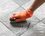 Mano con guante naranja cepillando un suelo antideslizante con espuma.