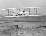 Wright Flyer mit Doppelruder hebt ab, Pilot liegt auf dem Flügel. Ein Mann beobachtet den historischen Flug.