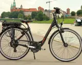Czarny rower elektryczny z baterią na bagażniku, gotowy do jazdy po bulwarach z widokiem na Wawel.