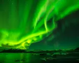 Grön norrsken dansar över en isig ö med stjärnor som glittrar. Vattnet speglar det magiska skådespelet.