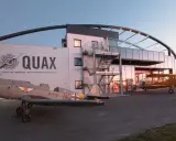 Zwei historische Flugzeuge stehen vor dem Quax Hangar. Ein graues Propellerflugzeug im Vordergrund, ein gelbes Doppeldeckerflugzeug daneben.