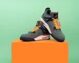 Jordan Motorsport Sneaker in Olivgrün mit orangefarbenen Schnürsenkeln, auf einem orangefarbenen Podest vor hellgrünem Hintergrund.