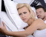 Donna bionda a letto con smartphone, sguardo preoccupato. L'uomo dorme accanto, forse ignaro del tradimento.
