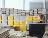 Frau sitzt vor einem Berg von Akten und Ordnern, die zeigen, wie wichtig es ist, bedienungsanleitungen aufbewahren.