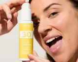 Une femme souriante tient un flacon de crème solaire visage SPF 30. La quantité de crème solaire est parfaite pour une protection optimale.