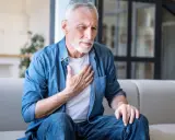Älterer Mann mit COPD hält sich die Brust. Er sitzt auf einem Sofa und sieht besorgt aus.