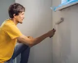Mężczyzna w żółtej koszulce maluje ścianę wałkiem. Czy przed każdym malowaniem trzeba gruntować ściany? To pytanie zadaje sobie wielu majsterkowiczów.