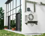 Unité extérieure d'une pompe à chaleur installée sur un mur blanc, à côté d'une maison moderne avec de grandes fenêtres. Comment choisir quelle pompe à chaleur choisir pour votre maison ?