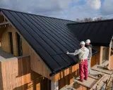 Zwei Handwerker sanieren ein Dach mit schwarzer Trapezblechdeckung. Das Holzhaus erhält ein modernes Aussehen.