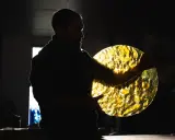 Artisan tenant un disque de verre texturé, illuminé par derrière. Les reflets dorés rappellent des vitraux.