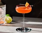 En vackert orangefärgad Division Bell cocktail med en apelsinzest som garnityr, bredvid en cocktailshaker och limeklyftor.
