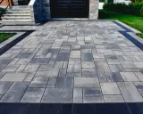 Nowoczesny podjazd z kostki brukowej w odcieniach szarości, z czarną obwódką. Wygląda jak piasek polimerowy, ale jest trwały i estetyczny.