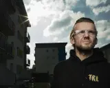 Mężczyzna w okularach patrzy w górę, na tle bloków mieszkalnych. Słońce przebija się przez chmury, tworząc efekt kękę dzieci.