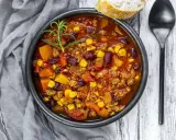 Herzhaftes Chili sin Carne mit Bohnen, Mais und Kürbis, garniert mit Rosmarin. Dazu ein Stück Brot und ein Löffel.