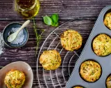 Goldbraune Zucchini-Muffins auf einem Gitter, einige in einer Muffinform. Zutaten wie Öl, Salz und Kräuter liegen bereit.