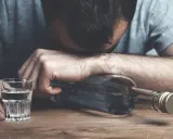 Mężczyzna z głową opartą o butelkę, obok kieliszek z alkoholem. Wygląda na to, że doświadcza zatrucia alkoholowego.