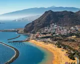 Bunte Häuser schmiegen sich an steile Hänge, während ein goldener Strand und das blaue Meer die Küste von Spanien zieren. Im Hintergrund erheben sich majestätische Berge.