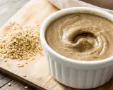 Un bol de sauce tahini crémeuse, à côté d'une pile de graines de sésame sur une planche en bois.