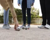 Une main de nom joueur de pétanque français saisit une boule argentée sur un terrain de gravier. D'autres boules et un cochonnet sont visibles.