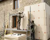 Deux ouvriers installent des panneaux isolants sur un mur en pierre. Une application concrète de l'isolation thermique, une définition essentielle pour l'efficacité énergétique.