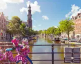 Das **beste von Amsterdam**: malerischer Kanal mit Booten, historischen Gebäuden und einem Kirchturm. Blumenverzierte Fahrräder auf einer Brücke.