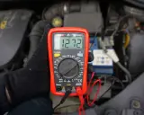 Ręka w czarnej rękawiczce trzyma multimetr pokazujący 12.72V, co pomaga sprawdzić akumulator.
